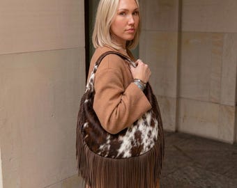 May include: A hobo-style shoulder bag featuring a brown and white cowhide pattern on the upper section and long, solid brown leather fringe detailing along the bottom edge. The bag has a single integrated shoulder strap and a curved silhouette.