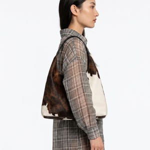 May include: A shoulder bag featuring a cowhide pattern with patches of dark brown and white fur. The bag has a structured silhouette with a single strap and a flat base, showcasing a natural animal print texture across the exterior surface.