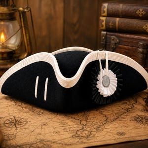 May include: A black tricorne hat featuring a white braided trim along the edges, two vertical white stripes on the side, and a circular black and white pleated cockade with a central silver-toned button accent attached to the front brim.