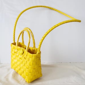 May include: A bright yellow woven basket bag featuring a rectangular base, two short rounded handles, and a long braided shoulder strap. The bag is constructed from a grid-like pattern of yellow strips, with a small circular closure at the top center.