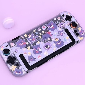 Puede incluir: Una consola de juegos portátil con una funda protectora de color morado claro decorada con varias ilustraciones del personaje Gengar, pequeñas estrellas, corazones e iconos de Poké Ball. El dispositivo incluye botones y joysticks negros.