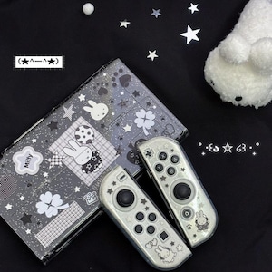 以下が含まれることがあります： グリッター仕上げの黒いゲーム機本体と2つのコントローラー。白いミッフィーのステッカー、クローバーのモチーフ、星、チェック柄で装飾されています。本体にはMiffyの文字が表示され、コントローラーには黒いボタンが付いています。