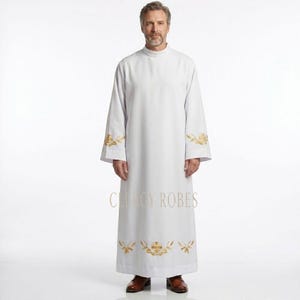 May include: A white clergy robe with gold embroidered accents on the sleeves and lower hem. The robe has a high collar and long sleeves. The text "CLERGY ROBES" is printed on the lower part of the robe.