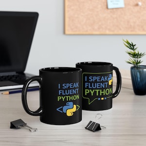 May include: Two black coffee mugs with the text "I Speak Fluent Python" in blue and green. One mug features a Python logo. The mugs are on a light-coloured surface with a laptop and office supplies in the background.