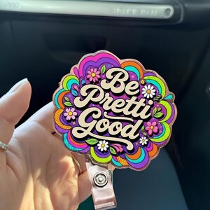 Pode incluir: Um porta-crachá colorido com as palavras "Be Pretti Good" em uma fonte retrô. O design apresenta um padrão floral e psicodélico em tons de roxo, laranja, verde e rosa. O porta-crachá tem uma alça de plástico transparente e um clipe de metal.