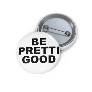 Puede incluir: Dos botones redondos blancos con respaldos de alfiler de metal. Un botón está boca arriba, mostrando el texto "BE PRETTI GOOD" en letras negras en negrita. El otro botón está boca abajo, mostrando el respaldo del alfiler de metal.