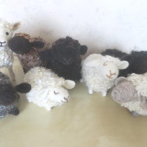 Könnte beinhalten: Eine Sammlung von sieben nadelgefilzten Schaf-Figuren in verschiedenen Farben, darunter Weiß, Schwarz, Grau und Braun. Jedes Schaf hat ein strukturiertes wolliges Fell, kleine Ohren und ausgeprägte Gesichtszüge und ist in einer Gruppe angeordnet.