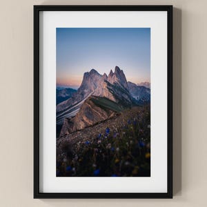 Puede incluir: Impresión enmarcada de un paisaje montañoso bajo un cielo azul claro. Los picos son escarpados y texturizados, con un degradado de colores de claro a oscuro. El primer plano muestra un campo de flores silvestres.