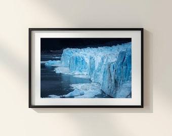 May include: A framed wall art print featuring a photograph of a massive, jagged glacier with bright blue ice formations meeting dark, calm water. The image is encased in a simple black frame with a wide white mat border, displaying a stark contrast between the icy landscape and the deep, shadowed background.