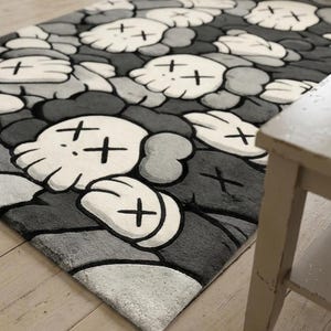 May include: A rectangular area rug featuring a repeating pattern of white cartoon-style skulls with crossed-out eyes and gloved hands, set against a background of overlapping dark gray and light gray rounded shapes with a soft, plush texture.