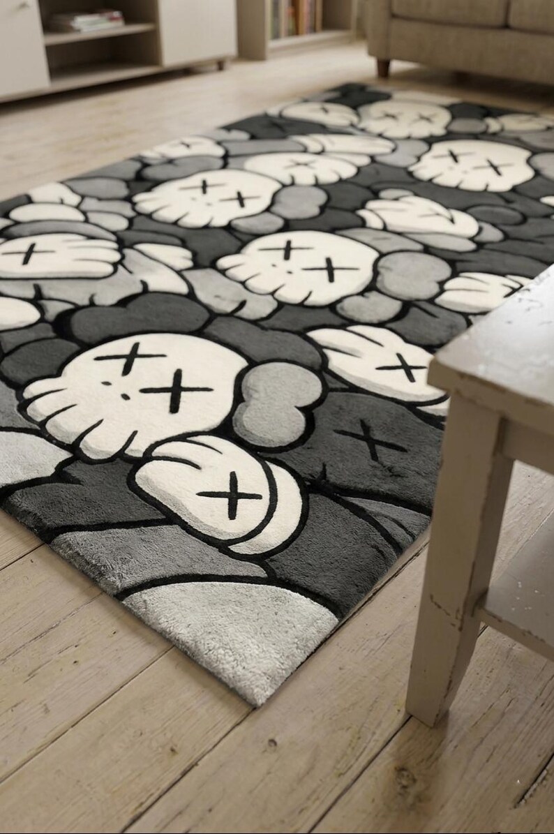 May include: A rectangular area rug featuring a repeating pattern of white cartoon-style skulls with crossed-out eyes and gloved hands, set against a background of overlapping dark gray and light gray rounded shapes with a soft, plush texture.