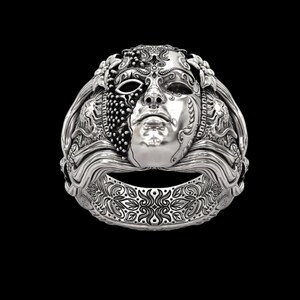 May include: A silver ring featuring a detailed mask design. The mask has a face with intricate details, including a half-covered eye and ornate patterns. The ring band also has a decorative floral design. The ring is set against a black background.