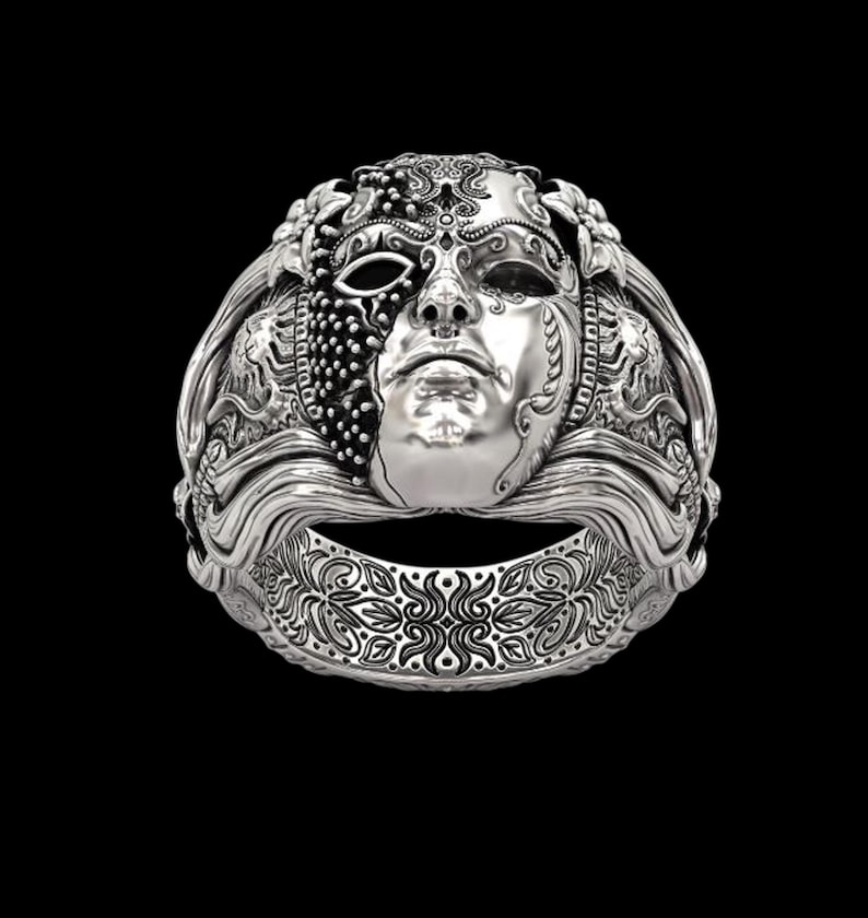 May include: A silver ring featuring a detailed mask design. The mask has a face with intricate details, including a half-covered eye and ornate patterns. The ring band also has a decorative floral design. The ring is set against a black background.