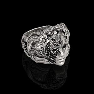 May include: A silver-toned ring featuring a detailed mask design. The mask is split, with one side adorned with floral and sun motifs, and the other with intricate patterns and a face. The ring has a reflective surface.
