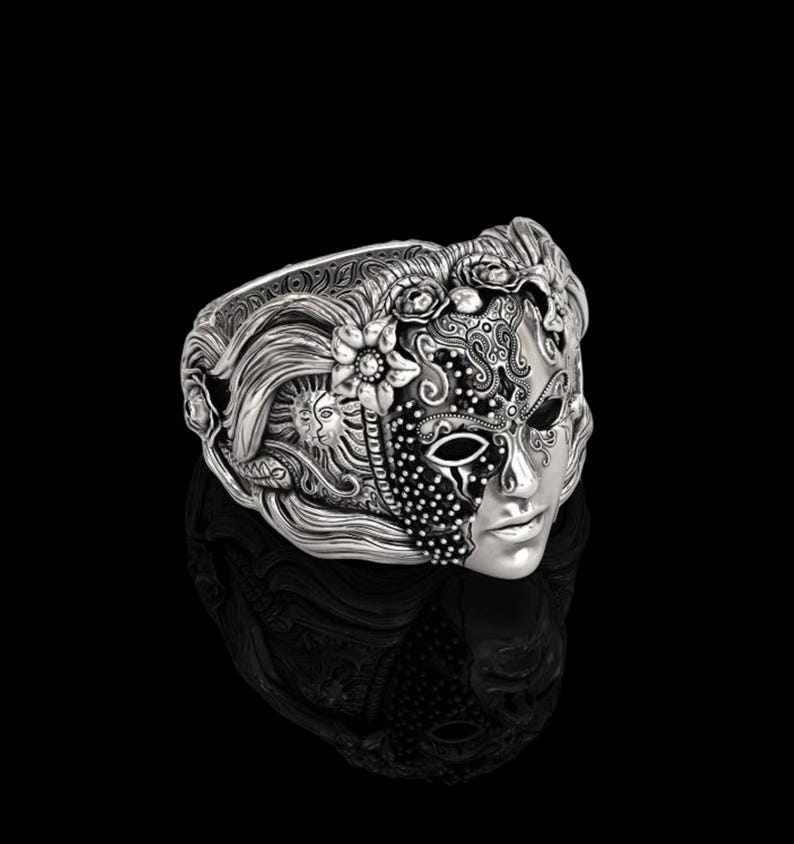 May include: A silver-toned ring featuring a detailed mask design. The mask is split, with one side adorned with floral and sun motifs, and the other with intricate patterns and a face. The ring has a reflective surface.