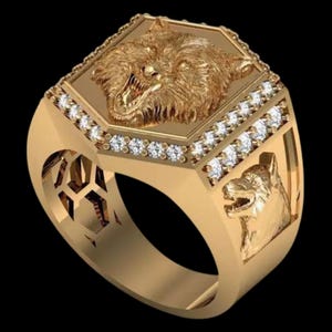 May include: A gold-colored ring featuring a detailed wolf head design. The ring has a rectangular face with a wolf's head and small, clear stones. The band also has a wolf head design on the side.