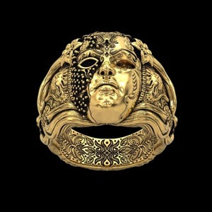 May include: A gold-colored ring with a detailed mask design. The mask features a half-face with intricate patterns and textures, including a beaded section and a smooth, sculpted face. The ring band has a floral design.