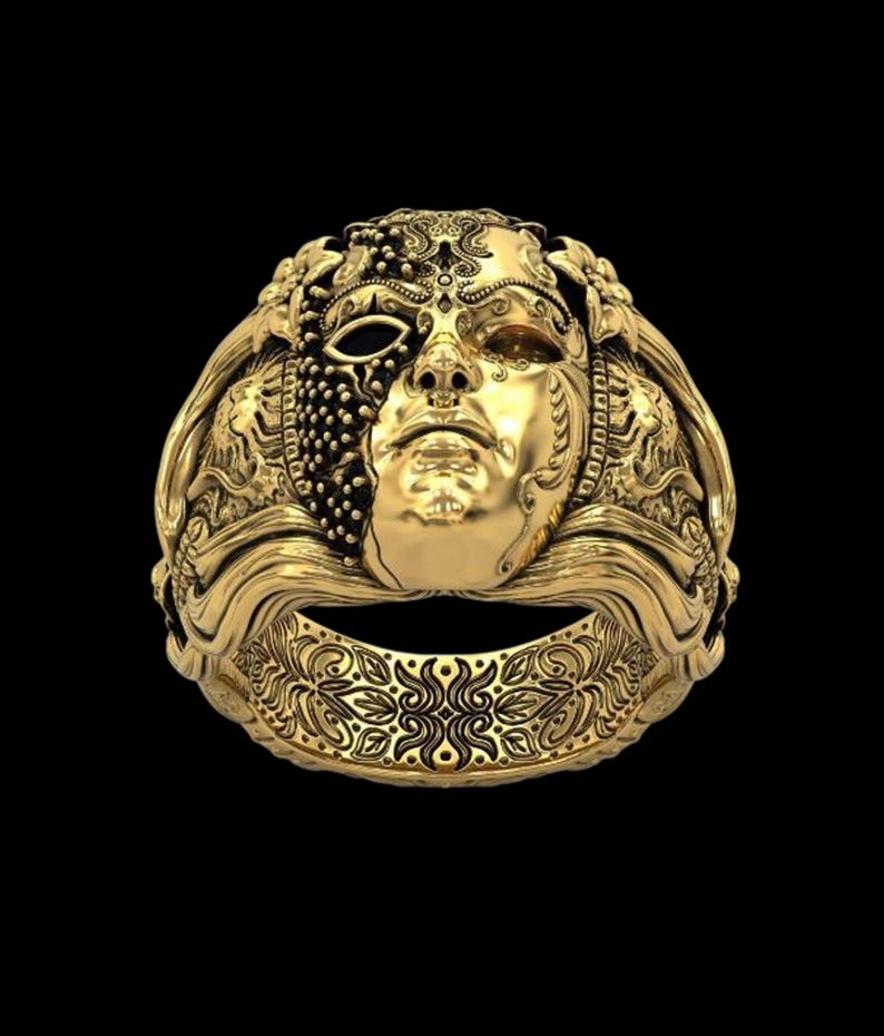 May include: A gold-colored ring with a detailed mask design. The mask features a half-face with intricate patterns and textures, including a beaded section and a smooth, sculpted face. The ring band has a floral design.
