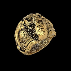 May include: A gold-colored ring featuring a detailed design of a face, a mask, and floral elements. The ring has intricate patterns and textures, with a dark background.