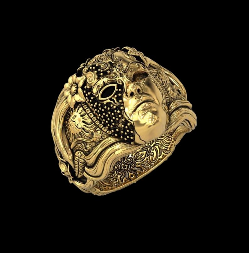 May include: A gold-colored ring featuring a detailed design of a face, a mask, and floral elements. The ring has intricate patterns and textures, with a dark background.