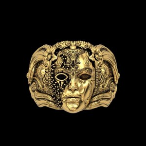 May include: A gold-colored ring featuring a detailed mask design. The mask is split, with one side adorned with intricate patterns and the other side depicting a face. The ring has a dark background.