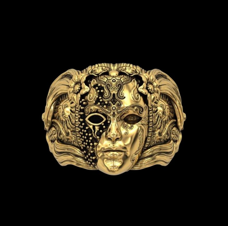 May include: A gold-colored ring featuring a detailed mask design. The mask is split, with one side adorned with intricate patterns and the other side depicting a face. The ring has a dark background.