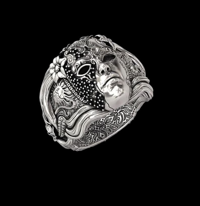 May include: A silver ring featuring a detailed mask design. The ring showcases a half-mask with intricate patterns, a floral element, and a sculpted face. The ring's design is highly detailed, with a polished finish.