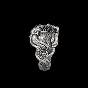 May include: A detailed silver ring featuring a woman's face with flowing hair, adorned with floral and sun motifs. The ring's design includes a rose, sun, and a dotted pattern, showcasing intricate craftsmanship. The ring is set against a black background.