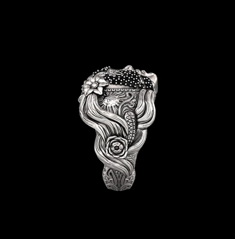 May include: A detailed silver ring featuring a woman's face with flowing hair, adorned with floral and sun motifs. The ring's design includes a rose, sun, and a dotted pattern, showcasing intricate craftsmanship. The ring is set against a black background.