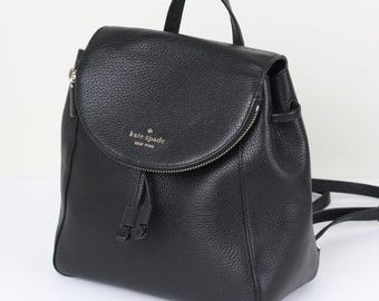 May include: A black pebbled leather backpack featuring a top handle, a fold-over flap with a zipper closure, and a drawstring detail. The front flap displays the gold-toned text kate spade NEW YORK. The bag includes adjustable shoulder straps.