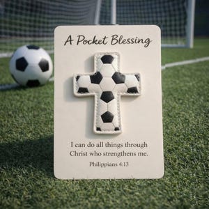 Pode incluir: Um cartão "A Pocket Blessing" com uma cruz de bola de futebol. A cruz é branca com um padrão de bola de futebol preta. O texto diz: "Tudo posso naquele que me fortalece. Filipenses 4:13". Uma bola de futebol no fundo.