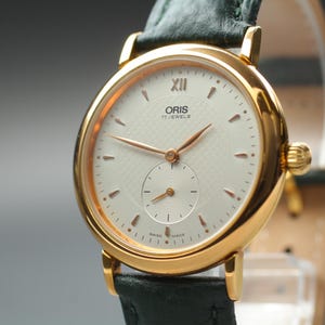 Może przedstawiać: Złoty zegarek Oris z białą tarczą i małą tarczą sekundową. Zegarek ma złote wskazówki i znaczniki oraz ciemnozielony skórzany pasek. Na tarczy widnieje napis "ORIS 17 JEWELS".