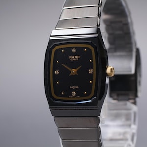 Puede incluir: Un reloj Rado Quartz Diastar negro con bisel y detalles dorados. La esfera del reloj es negra con agujas doradas e indicadores de hora de diamantes. La correa es de metal plateado.