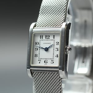 May include: A rectangular Longines watch with a silver mesh strap. The watch face is white with black numerals and hands. The watch has a classic design, suitable for everyday wear.