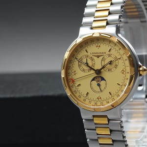 May include: A gold and silver-toned Longines watch with a light gold face. The watch features multiple subdials, a date display, and a moon phase indicator. The watch band is a combination of silver and gold links.