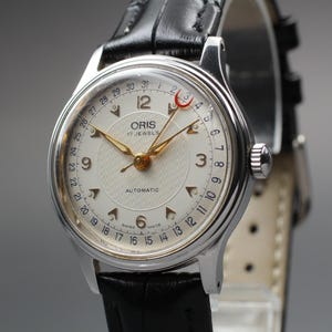May include: A vintage Oris automatic wristwatch with a silver case and a black leather strap. The cream-coloured watch face features gold hands and hour markers. The watch has a date display and the text "ORIS 17 JEWELS AUTOMATIC".