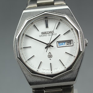 Puede incluir: Un reloj Seiko Grand Quartz plateado con esfera blanca y agujas negras. La esfera muestra el día como "SAB 29". El reloj tiene una correa plateada y una caja geométrica.