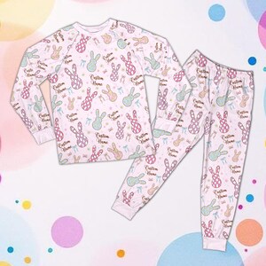 May include: A white pajama set with a repeating pattern of pastel-coloured bunnies and bows. The long-sleeved top and pants feature the text "Custom Name". The pajamas are made of a soft material.