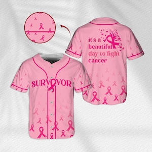 Op de afbeelding: Een lichtroze honkbalshirt met een knoopsluiting aan de voorzijde, korte mouwen en een herhalend patroon van donkerroze linten voor kankerbewustzijn. De voorzijde toont het woord SURVIVOR en de achterzijde it's a beautiful day to fight cancer.