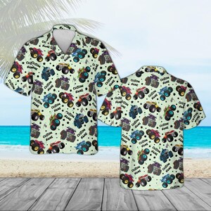 May include: Light green short-sleeve button-down shirt featuring a repeating pattern of colorful monster trucks and stars. The text "CUSTOM NAME" is printed on the shirt. The shirt is displayed on a wooden surface with a beach and ocean background.