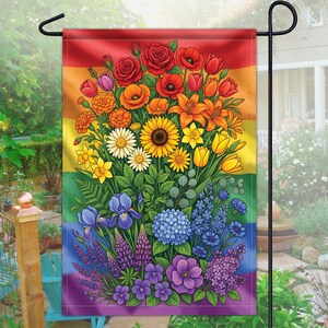 Puede incluir: Una bandera de jardín con un fondo de arcoíris y un colorido arreglo floral. El ramo incluye rosas rojas, lirios naranjas, girasoles amarillos e iris moradas. La bandera se muestra en un soporte de metal negro.