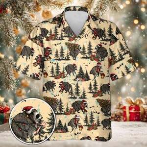 May include: A short-sleeve button-down shirt in a light beige color featuring an all-over print of raccoons wearing Santa hats and scarves, surrounded by pine trees, wrapped gift boxes, and string lights, with a circular inset detail.