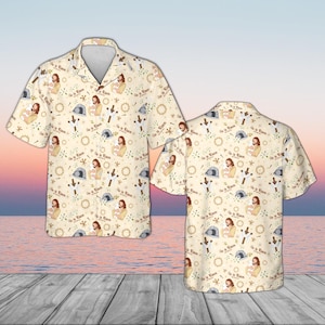 May include: A short-sleeved button-down shirt with a cream-colored background. The shirt features a repeating pattern of religious imagery, including a figure, a tomb, a cross, and the text "He is Risen".