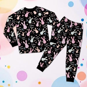 May include: Black pajama set with a repeating pattern of bunny rabbits in pink, blue, and yellow. The long-sleeved top and pants feature the text "Custom Name". The pajamas have black cuffs and a black crew neck.