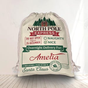May include: A cream-colored drawstring gift bag featuring a North Pole Express graphic with green pine trees, red text, and a gift box. The bag is printed with the name Amelia, a checkmark next to Nice, and a North Pole postmark with a sleigh icon.