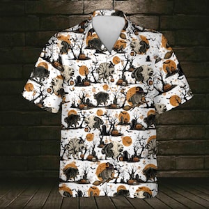 Op de afbeelding: Een overhemd met korte mouwen en knoopsluiting op een witte ondergrond met een all-over Halloween-print. Het patroon bevat wasberen in kostuums, pompoenlantaarns, kale bomen, grafstenen en oranje volle manen tegen een donkere achtergrond.