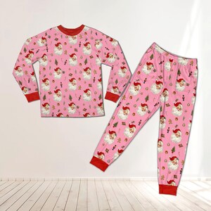 May include: A long-sleeve pajama set in light pink featuring an all-over print of Santa Claus faces, small green Christmas trees, and nutcracker figures. The shirt has a crew neckline, and both the shirt and pants have solid red ribbed cuffs.