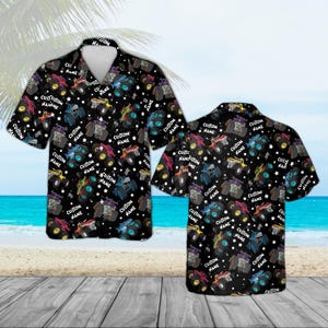 May include: Black button-down shirt with a repeating pattern of colorful monster trucks, stars, and the text "CUSTOM NAME". The short-sleeved shirt is displayed on a beach background.