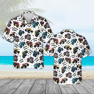 May include: White short-sleeve button-down shirt featuring a repeating pattern of colorful monster trucks, stars, and checkered flags. The text "CUSTOM NAME" is also printed on the shirt. The shirt is displayed on a wooden surface with a beach and ocean background.