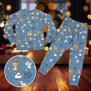 Puede incluir: Un conjunto de pijama azul de manga larga con un patrón repetitivo de figuras de la natividad, camellos, ovejas y ángeles. El estampado incluye el texto Glory to God y Holy Night entre estrellas amarillas y follaje verde.